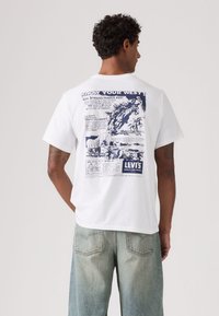 Λευκό βαμβακερό μπλουζάκι με κορυφαίο μπλε γραφικό σχεδιασμό στην πλάτη, που απεικονίζει εικονογραφήσεις και κείμενα με θέμα τη Δύση από τη Levi's.