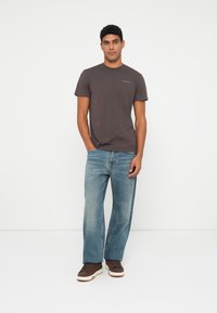 Un tânăr care stă cu mâinile în buzunare, purtând un tricou gri închis cu mânecă scurtă, blugi deschisi la culoare, cu croială largă, și adidași maron.