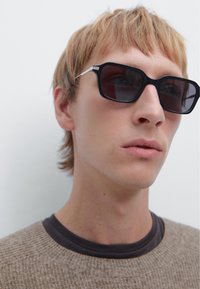 Black sunglasses with a geometric shape, dark lenses, and silver detailing on the temples. The frame has a glossy finish.