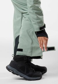 Water-resistant jacket in light green with black accents, featuring Velcro straps and a hand gripping the lower hem. Black hiking boots visible.