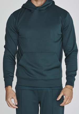Homme portant un sweat à capuche bleu sarcelle foncé avec une poche kangourou et un pantalon assorti, debout devant un fond uni.