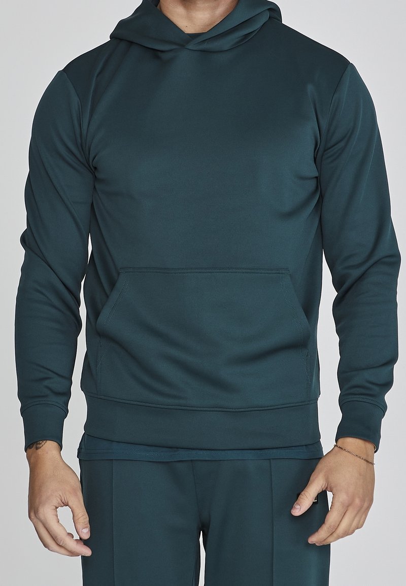 Homme portant un sweat à capuche bleu sarcelle foncé avec une poche kangourou et un pantalon assorti, debout devant un fond uni.