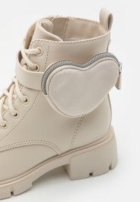 Stivaletto alla caviglia in pelle sintetica beige con punta tonda, suola spessa e taschino con zip a forma di cuore sul lato. Presenta dettagli di lacing e cuciture.