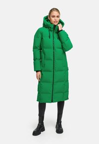 Groene pufferjas met een lange silhouet, ritssluiting en verstelbare capuchon. Heeft zijzakken en een gequiltde textuur. Draagt met zwarte jeans en laarzen.