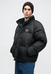 Μαύρο μπουφάν με ψηλό γιακά, που διαθέτει λογότυπο στο στήθος, φαρδιές πλάκες, ελαστικούς καρπούς και πλαϊνές τσέπες. Φοριέται με σκούφο σε σκούρο χρώμα.