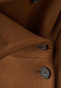 Bruin wollen stof met een diagonale weefstructuur, voorzien van een zwart knoopdetail en een omgevouwen kraag. Zichtbare stiksels aan de rand.