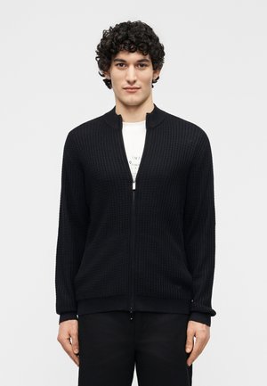 Tânăr cu păr creț, îmbrăcat într-un pulover negru cu fermoar și textură, peste un tricou alb cu imprimeu grafic, stând în fața unui fundal simplu.