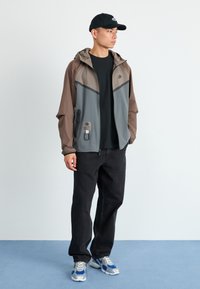 Veste à capuche grise et marron avec fermeture éclair à l'avant, poches et logo, associée à un t-shirt noir, un jean noir et des baskets avec des accents bleus.