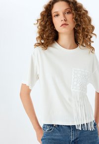 Camiseta blanca de algodón con un bolsillo de crochet texturizado que presenta detalles de flecos en el lado izquierdo, combinada con jeans de mezclilla azul.