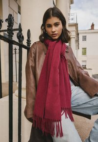Burgunderroter Schal aus weichem Material mit langen Fransen, drapiert über einer braunen Lederjacke, kombiniert mit hellblauen Jeans.