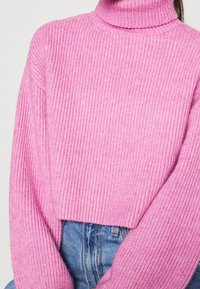 Pull en tricot rose avec un col montant, présentant une texture côtelée et un design court, associé à un jean en denim bleu.