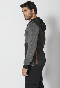 Sudadera con capucha en negro y gris, con un cuerpo texturizado, mangas en contraste y un detalle del logotipo naranja en el costado. Material de mezcla de algodón.