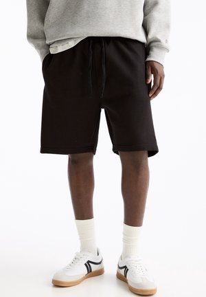 Man draagt zwarte short met trekkoord, witte sportsokken, witte sneakers met zwarte details en een grijze trui, met gedeeltelijk zichtbare handen.