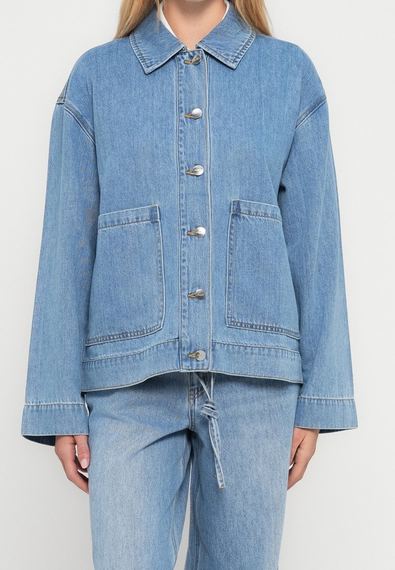 Femme portant une veste en jean bleu clair boutonnée avec deux poches avant et un pantalon en jean assorti sur fond blanc.