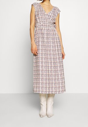 Kobieta w bezrękawnikowej sukience z dekoltem w serek, sięgającej do kostek, o wielokolorowym geometrycznym wzorze, ubrana w białe kowbojki, stojąca na jednolitym tle.