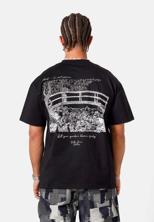 Mann mit geflochtenem Haar trägt ein schwarzes T-Shirt, das auf der Rückseite eine weiße Linienzeichnung einer japanischen Gartenbrücke und floraler Pflanzen zeigt.