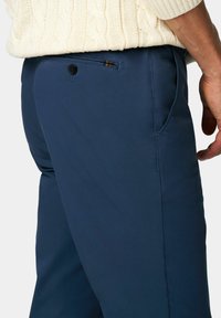 Marineblauwe broek met een gladde textuur, voorzien van een achterzak met een zwarte knoop en een subtiel geruit patroon op de voering van de zak.