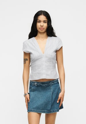 Nő hosszú sötét hajjal, fehér rövid ujjú V-nyakú felsőt és kék farmer szoknyát övvel, sima háttér előtt áll.