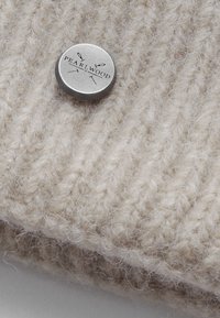 Šviesiai ruda megzta vilnos kepurė su raštuota tekstūra, turinti sidabrinį metalinį mygtuką, ant kurio išgraviruotas užrašas "PEARLWOOD". Minkšta ir šilta išvaizda.