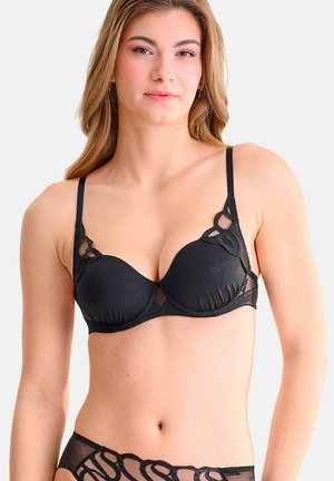 Jeune femme aux longs cheveux châtain clair portant un soutien-gorge en dentelle noire et une lingerie transparente assortie, debout devant un fond uni.