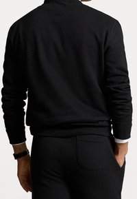 Svart sweatshirt med rundad halsringning och långa ärmar, med en slät textur. Matchas med svarta byxor och en synlig klocka.