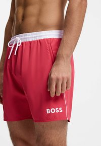 Άνδρας που φοράει κόκκινα μαγιό με λευκή ελαστική μέση, κορδόνι, πλαϊνές τσέπες και λογότυπο "BOSS" στο αριστερό πόδι.