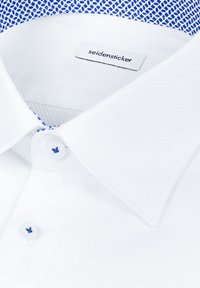 Chemise blanche boutonnée avec un tissu texturé, col mandarin, détail bleu à motifs à l'intérieur du col avec une étiquette de marque.