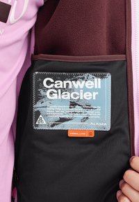 Svart innerficka med en blå etikett som säger "Canwell Glacier", en orange etikett "THERMAL LAYER 2" och vinröd fleecefoder. Rosa tyg i bakgrunden.