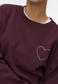 Persoon in een bordeauxrode sweatshirt met een strass hartontwerp op de borst, over een witte onderhemd, te zien van nek tot midden van de torso.