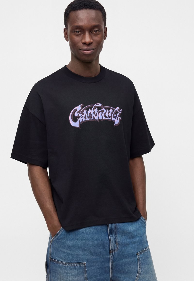 Junger Mann trägt ein schwarzes Carhartt T-Shirt mit lila Logo und blauen Jeans, steht mit den Händen in den Taschen vor einem schlichten Hintergrund.