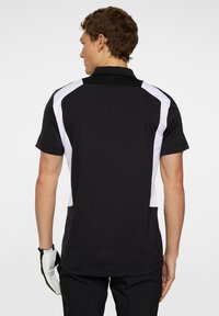 Svart golftröja med vita sidopaneler, korta ärmar och krage. Baksidan har ett texturerat område nära nacken. Texturerad tyg.