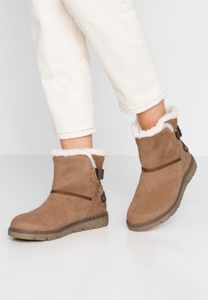 Benedenlichaam met beige broek en lichtbruine suède enkellaarsjes met witte bontvoering en details van een donkerbruine zool en gesp.