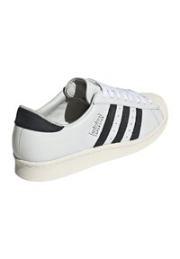 Sneaker in pelle bianca con strisce nere, suola in gomma e dettagli traforati. Presenta una punta arrotondata e parte posteriore traforata. Testo: "adidas SUPERSTAR."