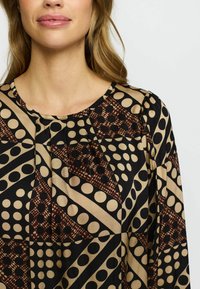 Kvinna som bär en blus med lång ärm, rund hals och geometriskt mönster med beige, svart och bruna prickar och former.