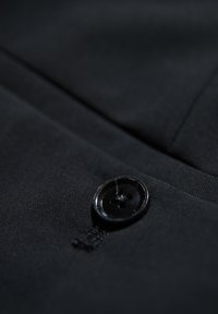 Zwarte stof met een gladde textuur, voorzien van een enkele ronde zwarte knop met vier gaten, waarbij de geïntegreerde naden zichtbaar zijn.