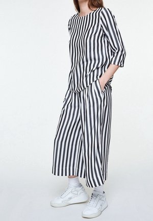 Frau trägt locker geschnittenes schwarz-weiß gestreiftes Oberteil und weitbeinige verkürzte Hose mit weißen Turnschuhen und Socken, steht mit Händen in den Taschen.