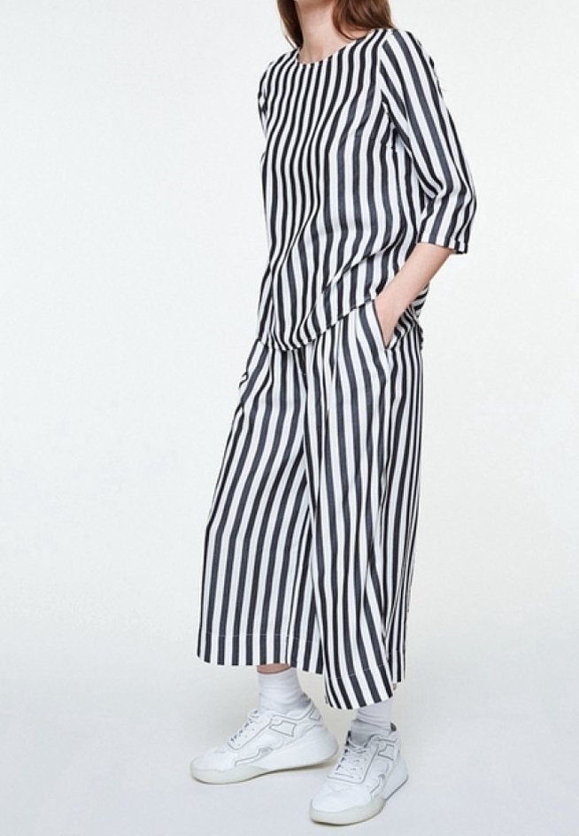 Femme portant un haut ample rayé vertical noir et blanc et un pantalon large court avec des baskets blanches et des chaussettes, debout les mains dans les poches.
