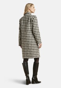 Zwart-witte lang jack met pied-de-poule-patroon, een getailleerd ontwerp, hoge kraag en zijzakken, gecombineerd met zwarte kniehoge laarzen met hak.