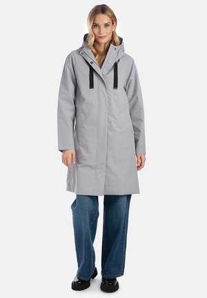 Vrouw die een lichtgrijze lange regenjas met capuchon, zwarte trekkoorden, blauwe spijkerbroek en zwarte laarzen draagt, staand tegen een witte achtergrond.