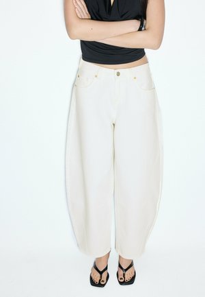 Person iført hvidtaljede, vidtbenede bukser, med armene krydset iført sort ærmeløs top og sorte åbne hælede sandaler.