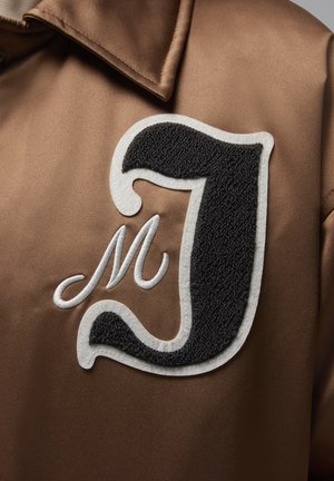 Krusiv brown satin jacket koja uključuje izvezeno crno slovo "J" s belim obrubima, kao i dodatni stilizovani beli detalj "M".