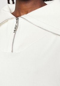 Witte sweater met rits en geribbelde textuur, grote capuchon en een opvallende zilveren ritssluiting met "HUGO" gegraveerd.