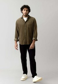Olivenfarvet kordfløjlsskjorte med en lomme, over en hvid t-shirt, parret med sorte bukser og hvide sneakers. Modellen står foran en grå baggrund.