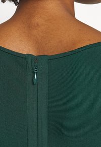 Haut en tissu texturé vert avec une fermeture éclair au dos dotée d'une tirette en teal. L'encolure est large et a une finition lisse.