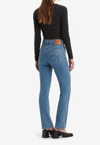 Högmidjade blå denimjeans med bakfickor dekorerade med rhinestones, kombinerade med en svart topp med långa ärmar och svarta klackskor.