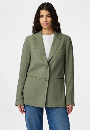 Vrouw met lang donker haar, gekleed in een licht olijfgroene blazer over een wit shirt en blauwe spijkerbroek, staand tegen een effen achtergrond.