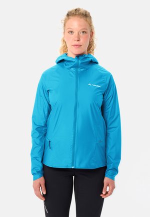 Frau mit lockigem Haar, die eine blaue Vaude-Jacke mit Kapuze und schwarze Hose trägt, stehend vor einem weißen Hintergrund.