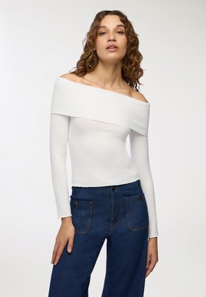 Top bianco a spalle scoperte in maglia a coste con maniche lunghe, abbinato a pantaloni in denim a gamba larga blu scuro con tasche frontali e dettagli in bronzo.