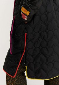 Čierna prešívaná bunda srdiečkovými vzormi, jasne farebnými obšitím v ružovej, oranžovej a žltej farbe, s pripojenými štítkami v štýle lístkov na rukáve.