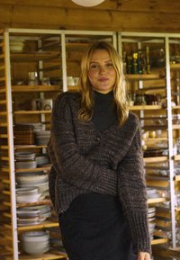 Sötét szürke kötött kardigán bordázott mintával és túlméretezett szabással, egy gombbal záródik és széles ujjakkal rendelkezik, fekete garbó fölött viselve.